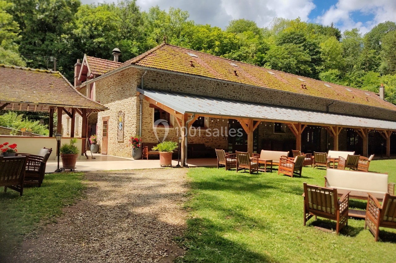 Bâtiment en pierre avec toit en tuiles, terrasse couverte et mobilier de jardin, entouré de verdure.