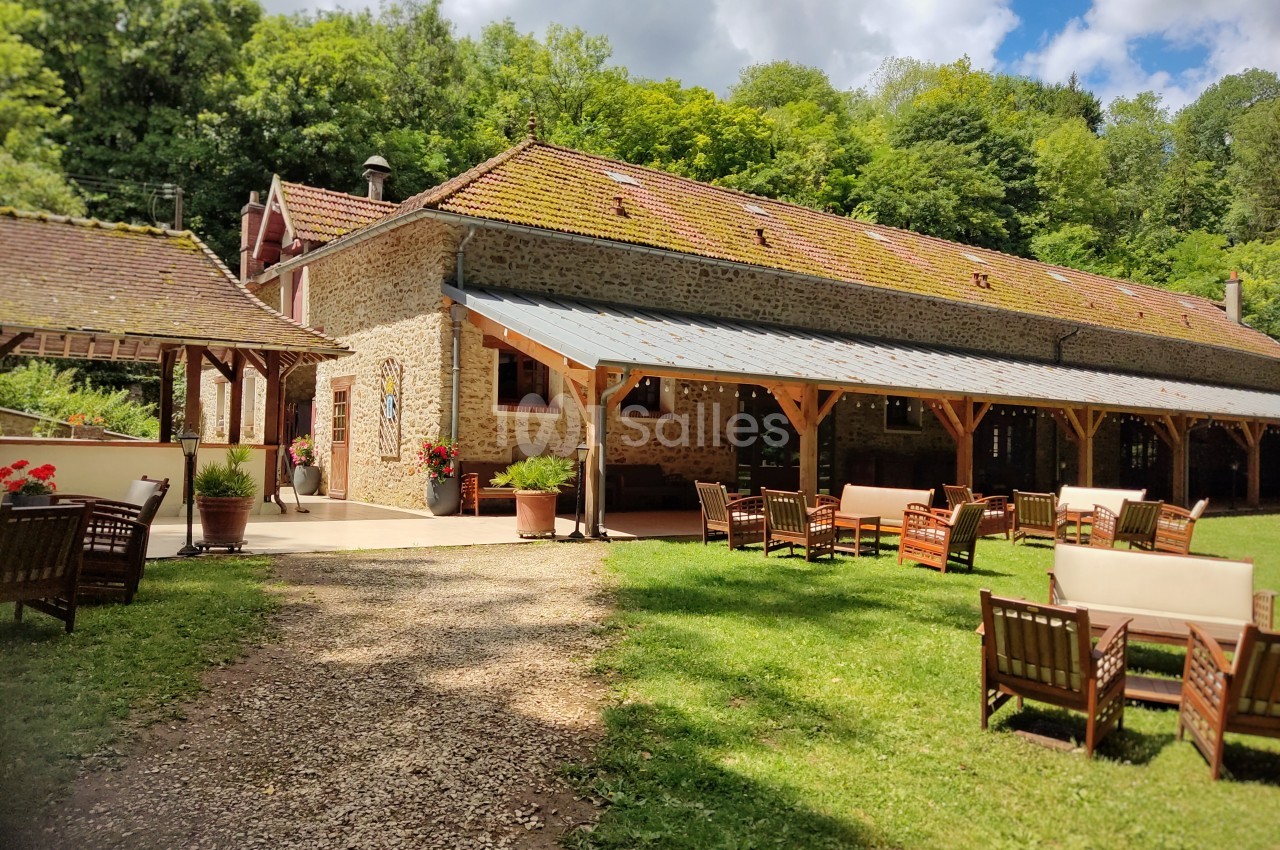 Bâtiment en pierre avec toit en tuiles, terrasse couverte et mobilier de jardin, entouré de verdure.