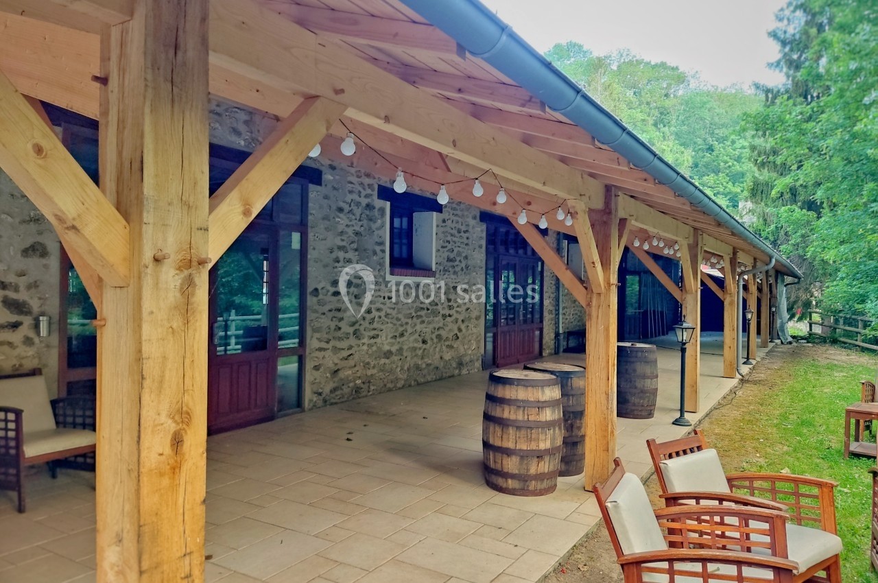 Terrasse couverte en bois avec des tonneaux décoratifs, des fauteuils et un mur en pierre, entourée de verdure.