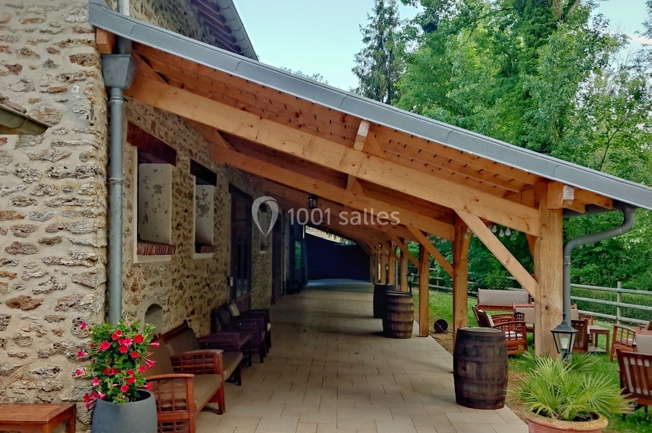 Terrasse couverte en bois attenante à un bâtiment en pierre, avec des tables, des chaises et des plantes en pot.