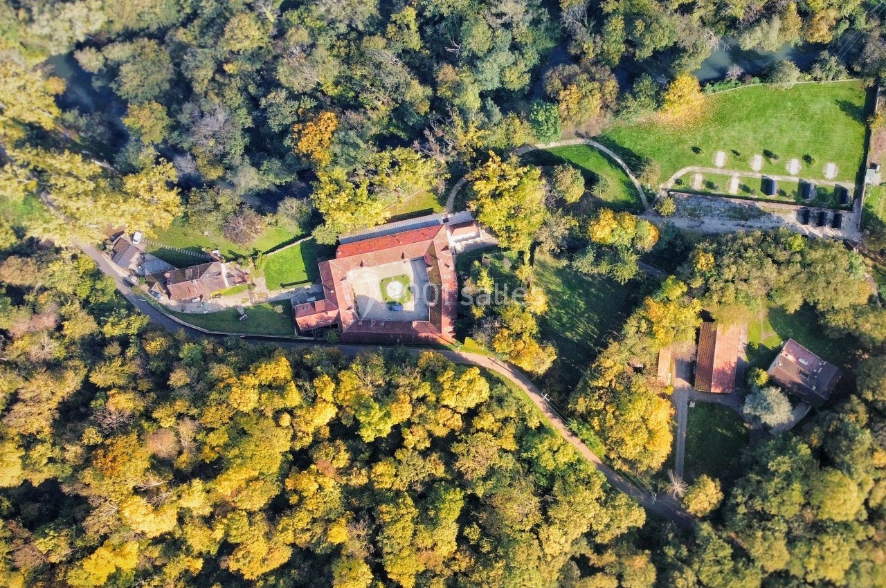 Vue aérienne d'un bâtiment entouré de forêt dense et de prairies, avec des chemins visibles autour.