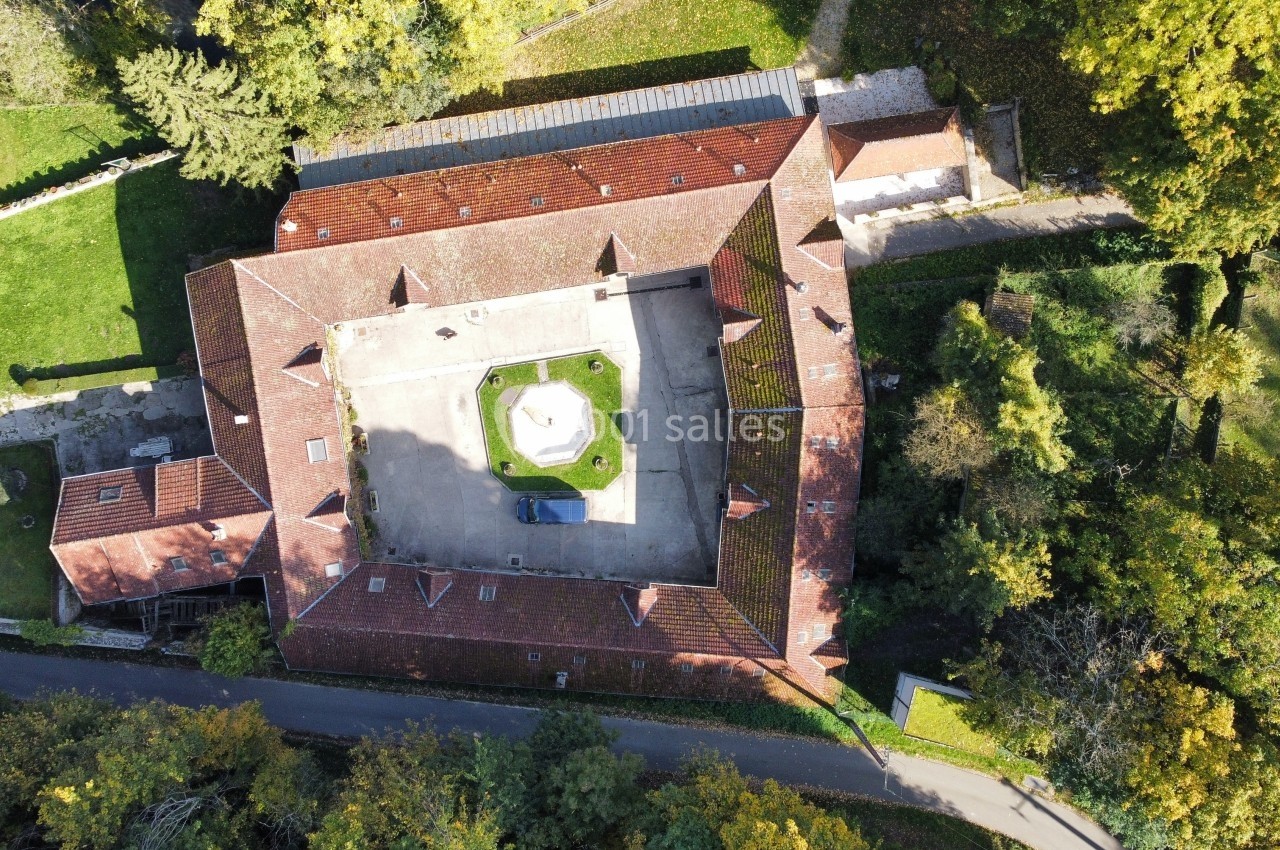 Vue aérienne d'un bâtiment historique en forme de carré avec une cour centrale, entouré de végétation et d'une route.