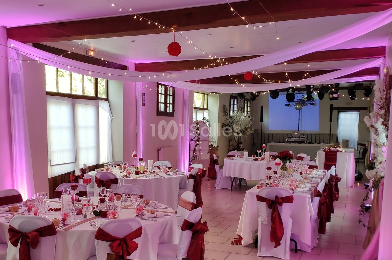 Salle de réception décorée avec des nappes blanches, des rubans rouges, des guirlandes lumineuses et des tables dressées.