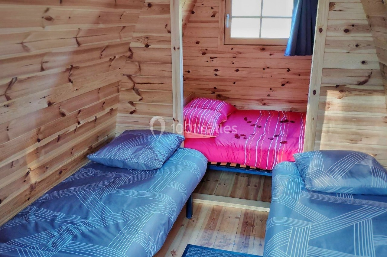 Deux lits simples avec draps bleus et un lit double avec draps roses dans une chambre en bois clair avec une fenêtre.