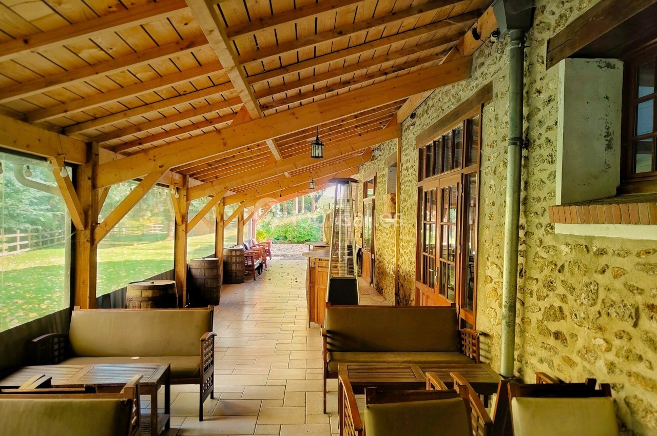 Terrasse couverte en bois avec mobilier de jardin, jouxtant un bâtiment en pierre et donnant sur un espace vert.