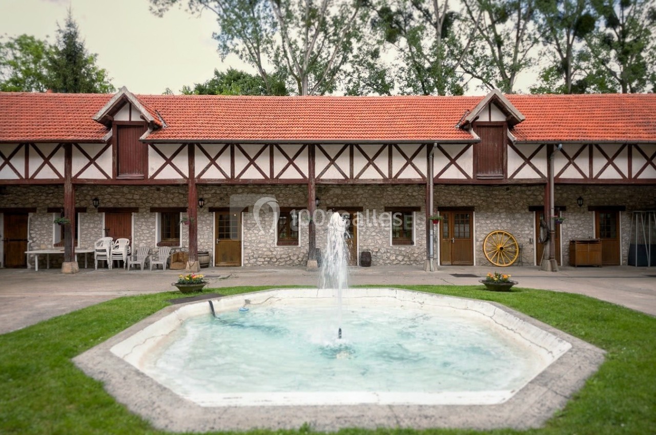 Cour intérieure avec une fontaine centrale, entourée de bâtiments en pierre et bois à toit rouge, bordée de verdure.