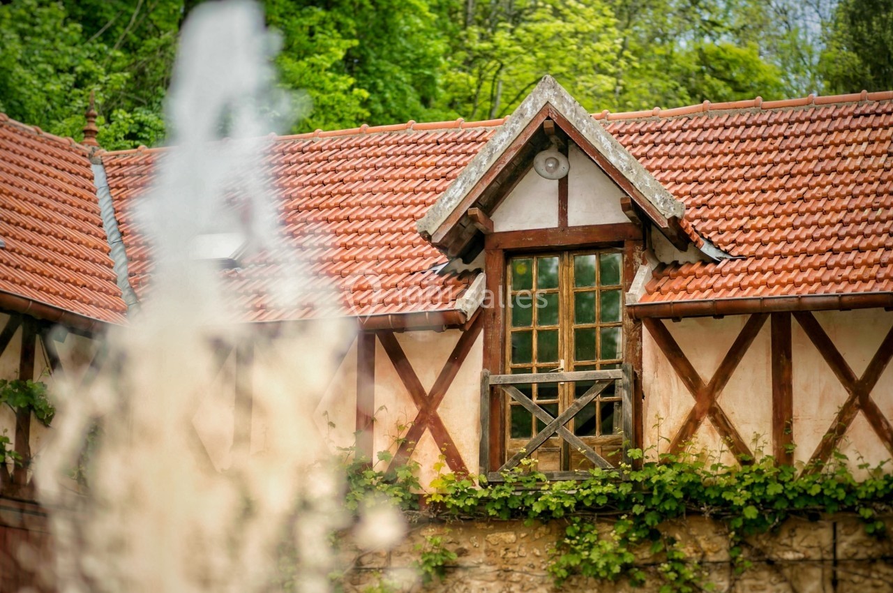 Façade d'une maison à colombages avec toit en tuiles rouges, entourée de végétation, vue partiellement derrière une fontaine.