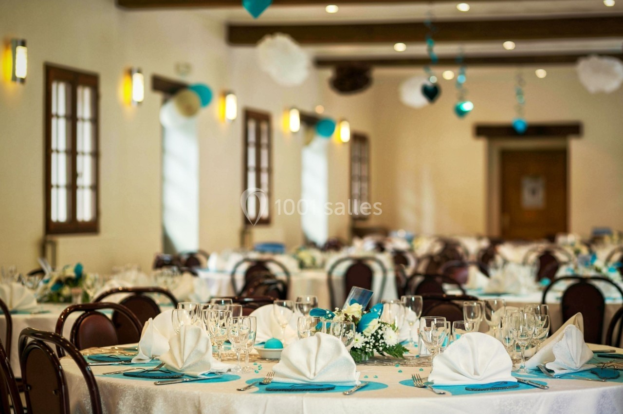 Salle de réception décorée avec des tables rondes dressées, ornées de nappes blanches et de décorations bleu et blanc.