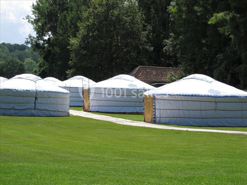 Des yourtes blanches alignées sur une pelouse verte, entourées d'arbres et d'un bâtiment en arrière-plan.