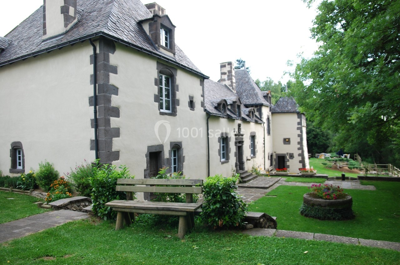 Façade d'une maison en pierre avec toit en ardoise, entourée d'un jardin verdoyant et d'un banc en bois.