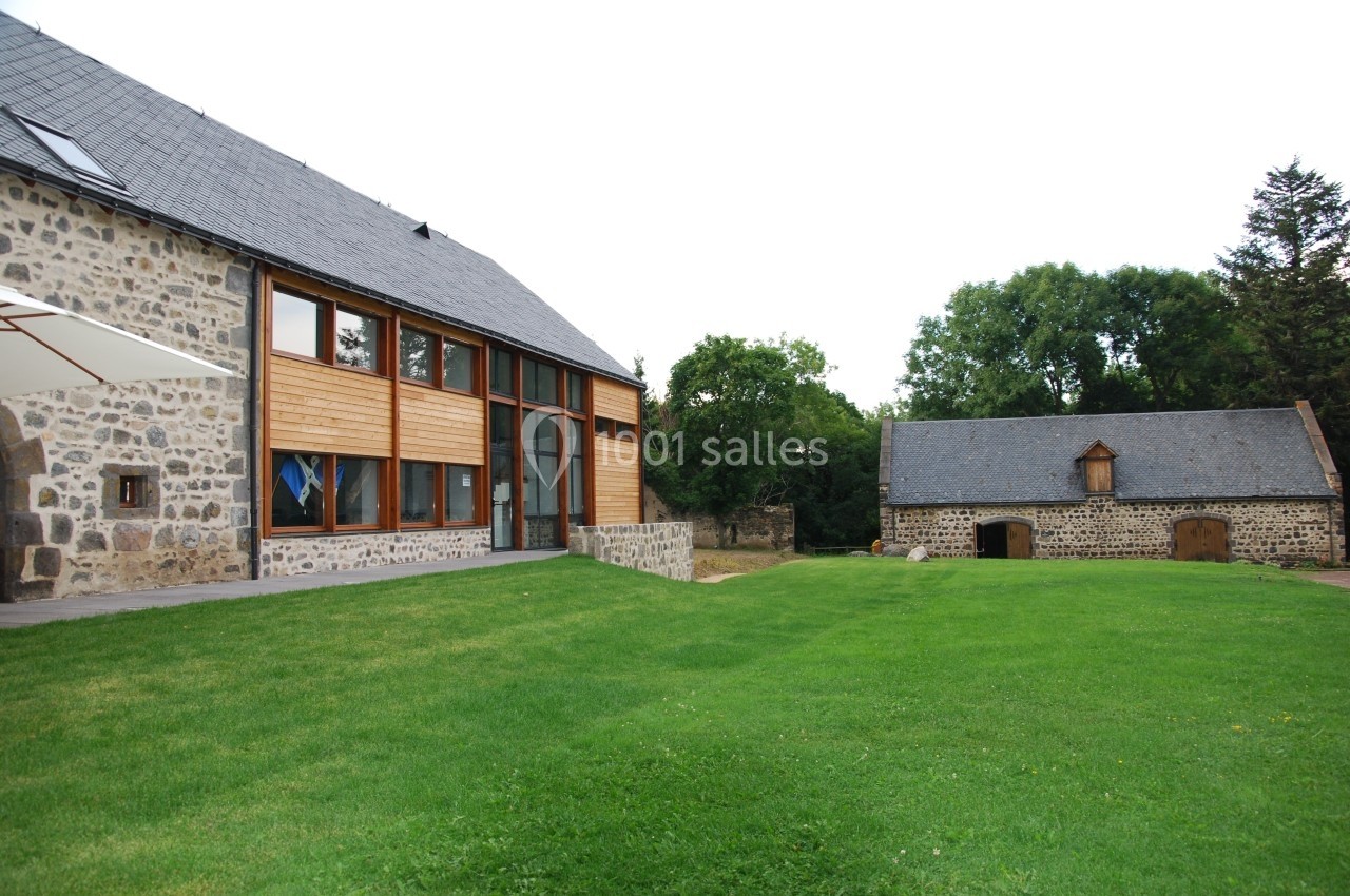 Bâtiment moderne en pierre et bois à gauche, petite grange traditionnelle à droite, entourés d'une pelouse verte.