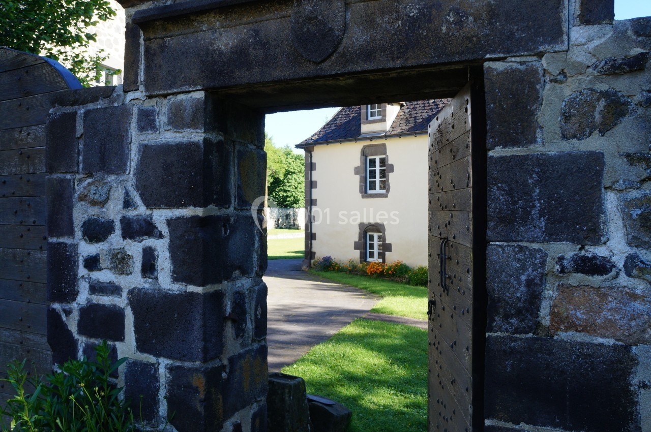 Portail en pierre ouvert donnant sur une maison en pierre avec des volets blancs et un jardin fleuri.