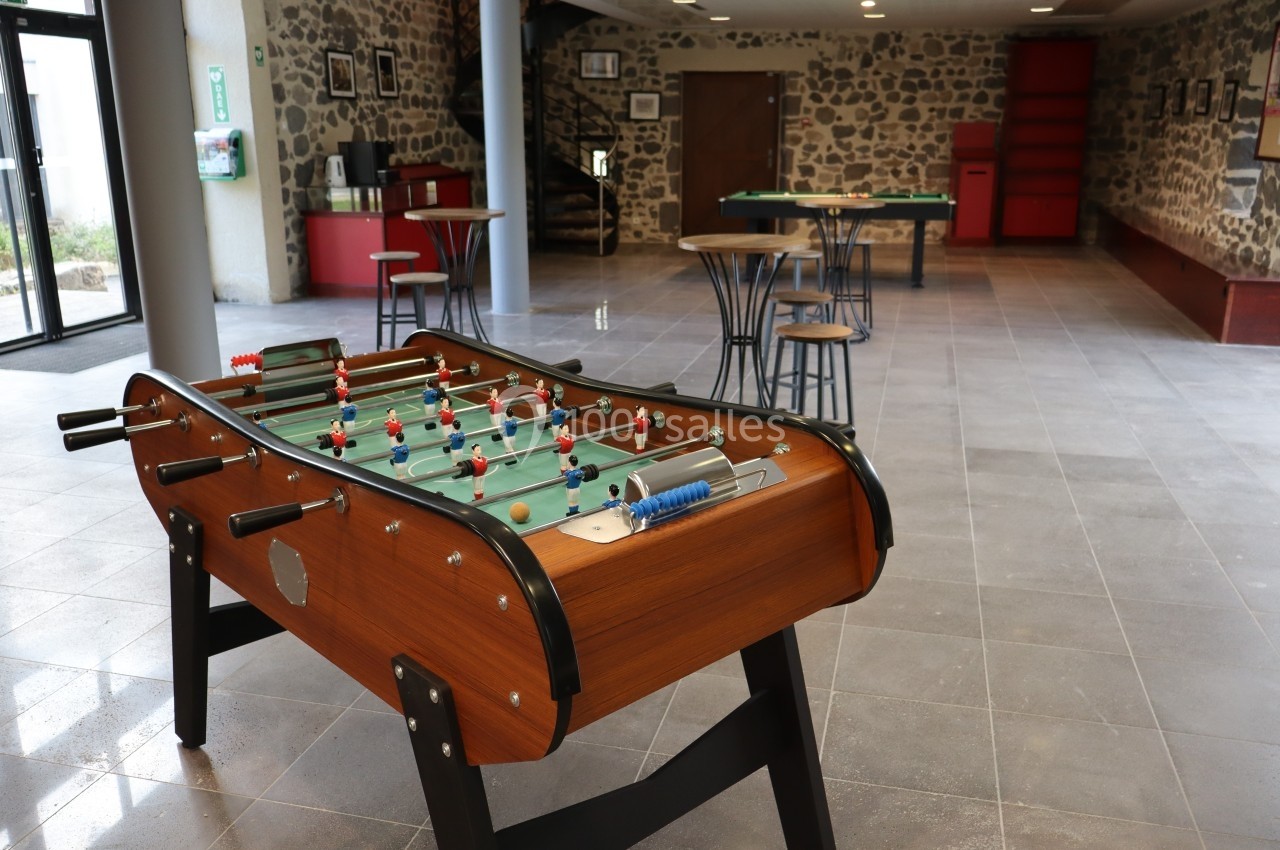 Baby-foot en bois dans une salle spacieuse avec tables, chaises et murs en pierre apparente.