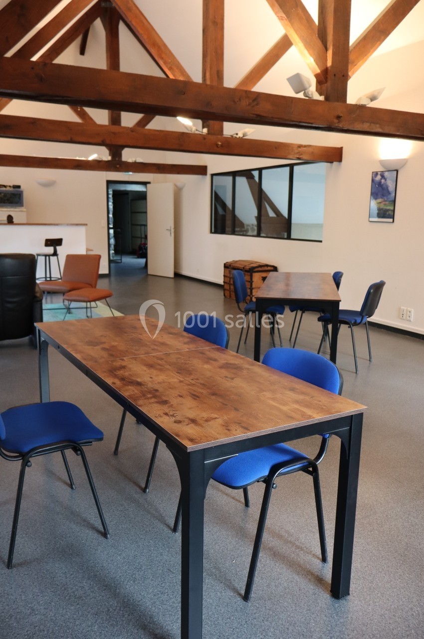 Salle lumineuse avec poutres apparentes, tables en bois, chaises bleues et espace ouvert en arrière-plan.