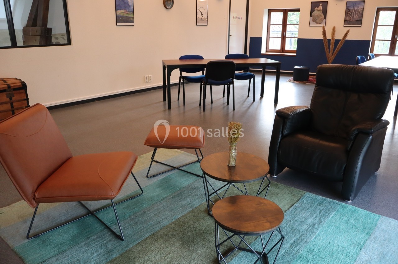 Espace salon avec fauteuils, tables basses et tapis, dans une pièce lumineuse avec des fenêtres et une table à l'arrière.