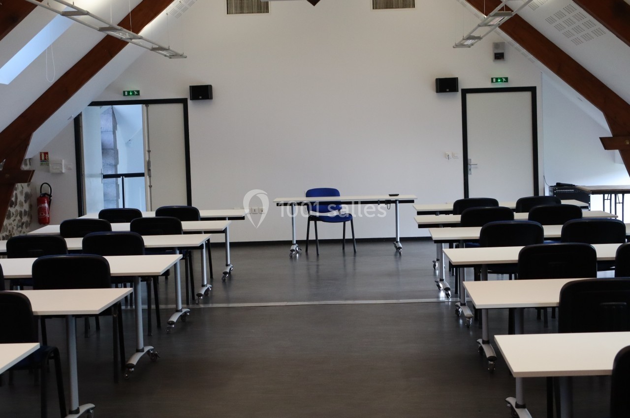 Salle de formation vide avec des tables blanches, des chaises noires et un bureau central sous un plafond en poutres…