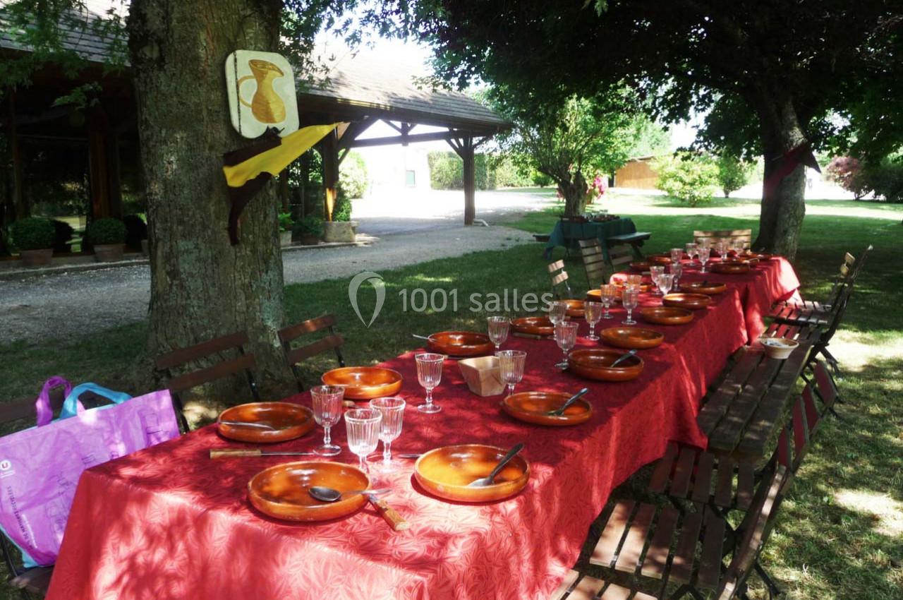 Table dressée en extérieur sous des arbres, avec nappes rouges, assiettes en terre cuite et couverts disposés.