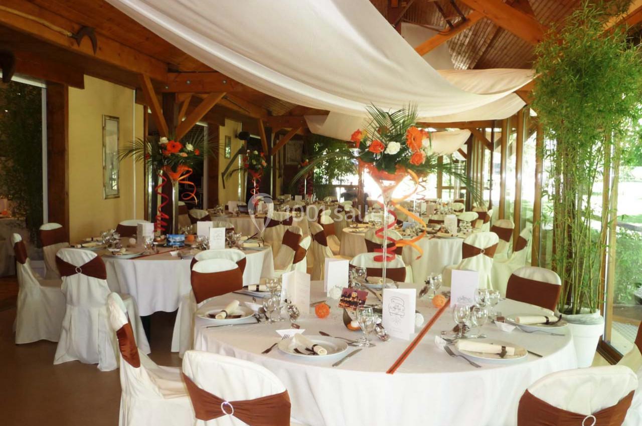Salle de réception décorée avec des tables rondes, nappes blanches, chaises ornées de rubans marron et compositions florales.