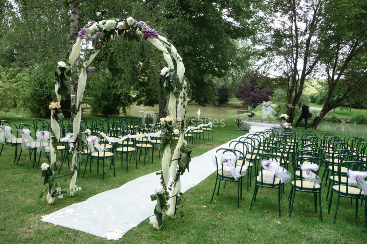 Allée centrale bordée de chaises décorées et arche fleurie dans un jardin verdoyant, préparée pour une cérémonie.