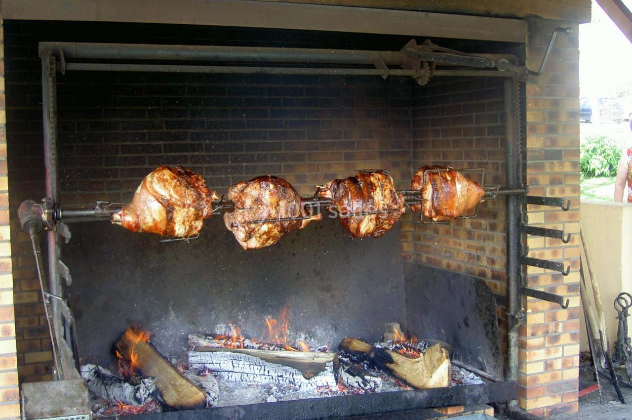 Quatre morceaux de viande rôtissent sur une broche au-dessus d'un feu de bois dans un foyer en briques.