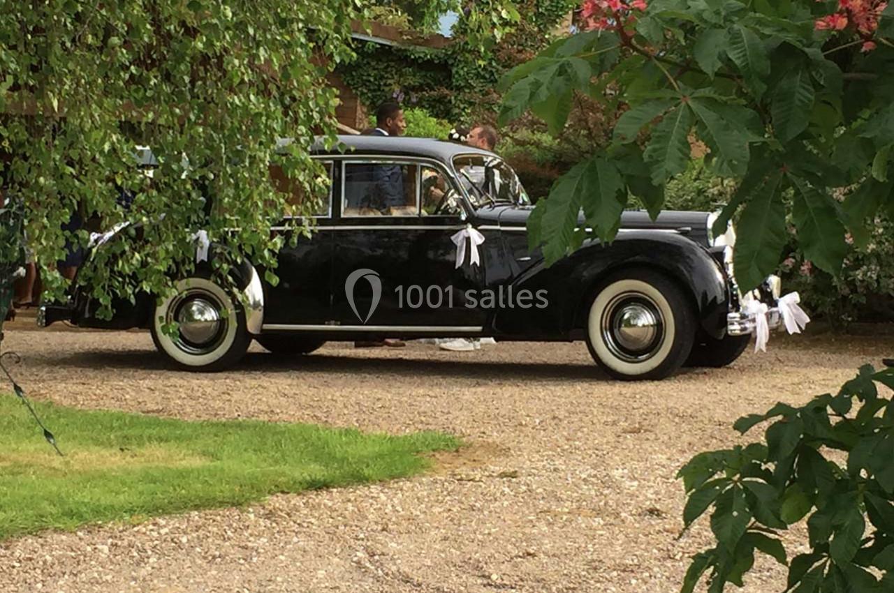 Voiture ancienne noire décorée de rubans blancs, stationnée sur une allée gravillonnée entourée de verdure.