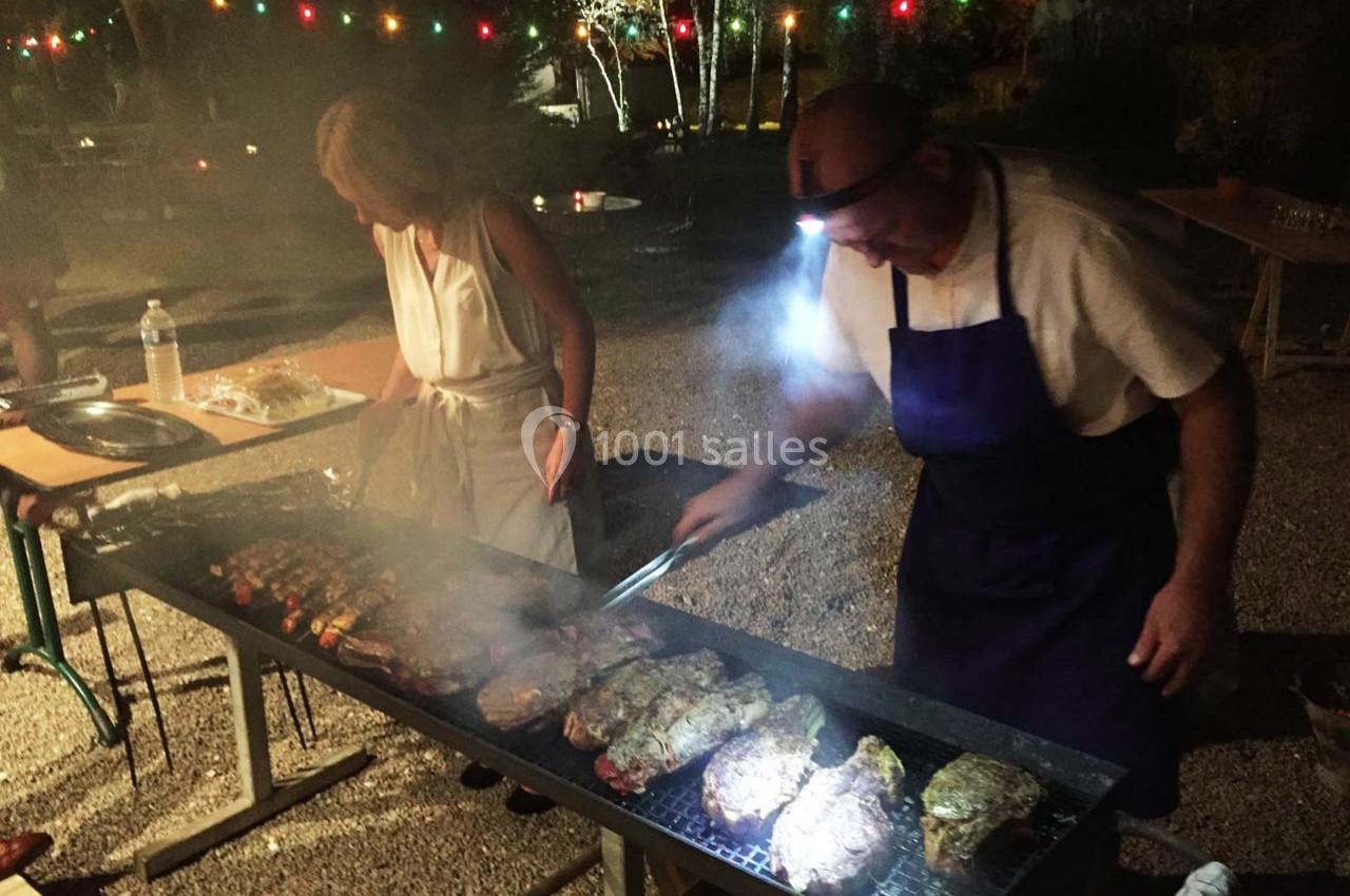 Deux personnes cuisinent des viandes sur un grand barbecue en extérieur, éclairé par des guirlandes lumineuses.