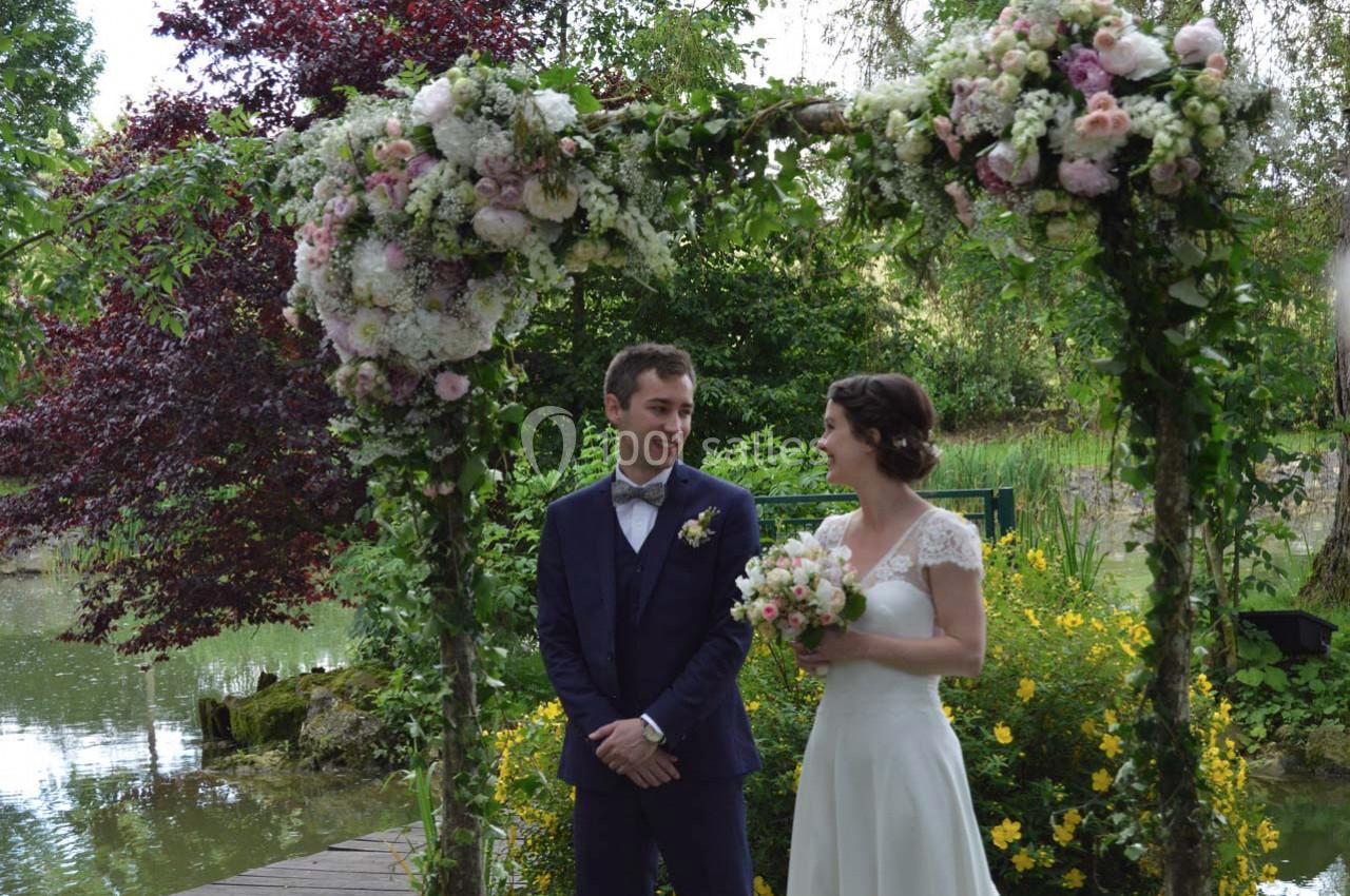Un couple en tenue de mariage se tient sous une arche fleurie dans un jardin près d'un étang.