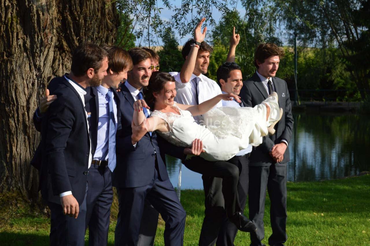 Un groupe d'hommes en costume porte une femme en robe de mariée dans un parc verdoyant près d'un plan d'eau.