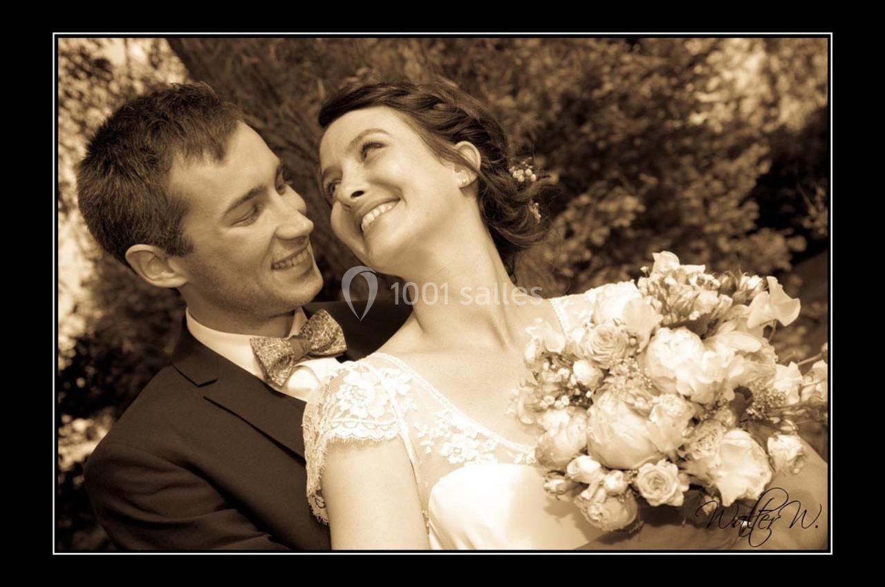 Un couple souriant en tenue de mariage, la mariée tenant un bouquet, dans un cadre extérieur arboré.
