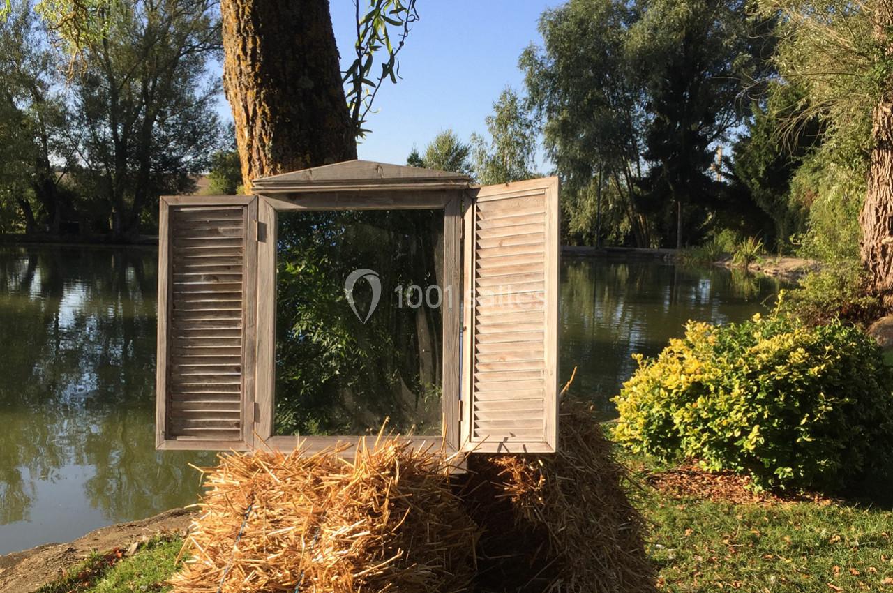 Miroir encadré de volets en bois, posé sur une botte de paille près d'un étang entouré de végétation.