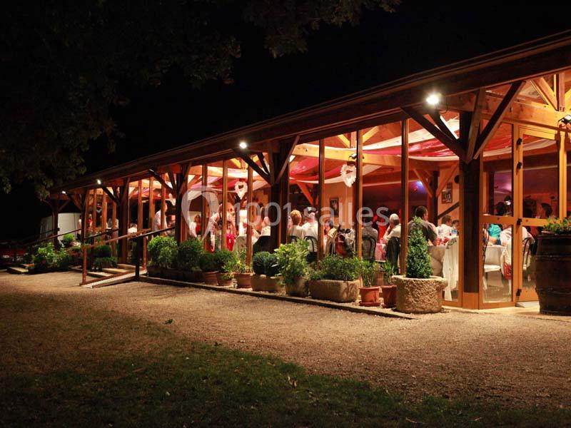 Vue nocturne d'une salle de réception vitrée éclairée, entourée de plantes et de graviers.