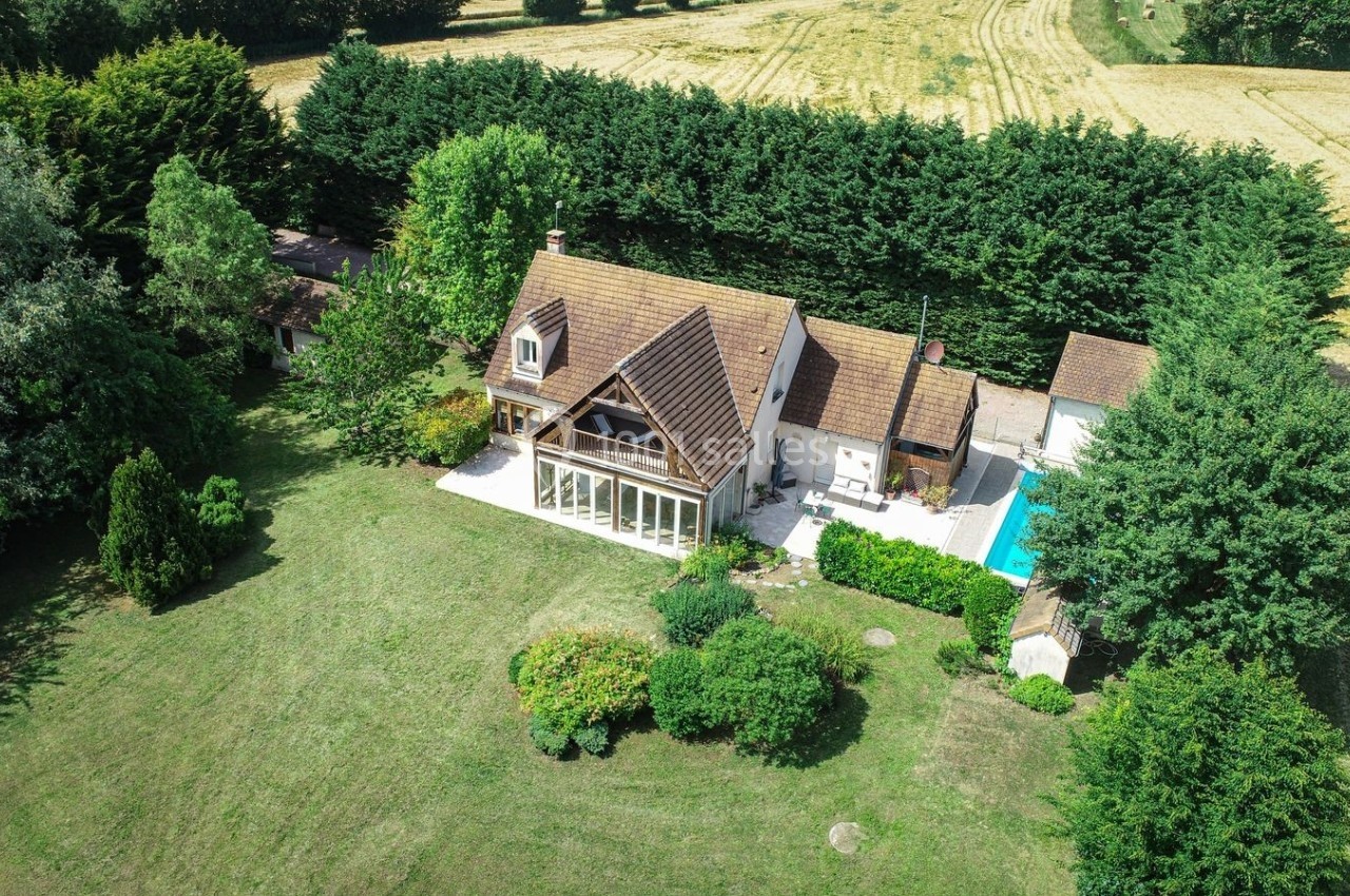 Vue aérienne d'une maison avec toit en pente, jardin arboré, piscine et champs environnants.