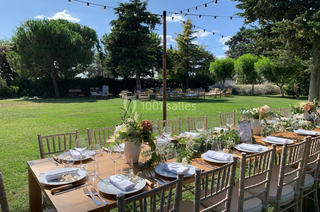 Location salle Pomérols (Hérault) - Domaine Fon de Rey #45 Table en bois décorée pour un repas en plein air, entourée de chaises, dans un jardin avec des arbres et guirlandes…