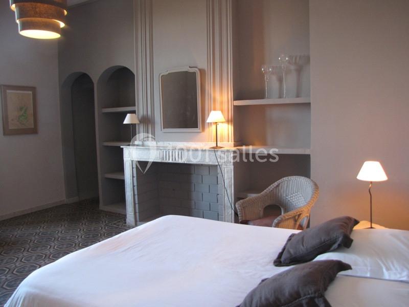 Location salle Pomérols (Hérault) - Domaine Fon de Rey #16 Chambre avec lit double, cheminée décorative, étagères intégrées, fauteuil en osier et éclairage doux.