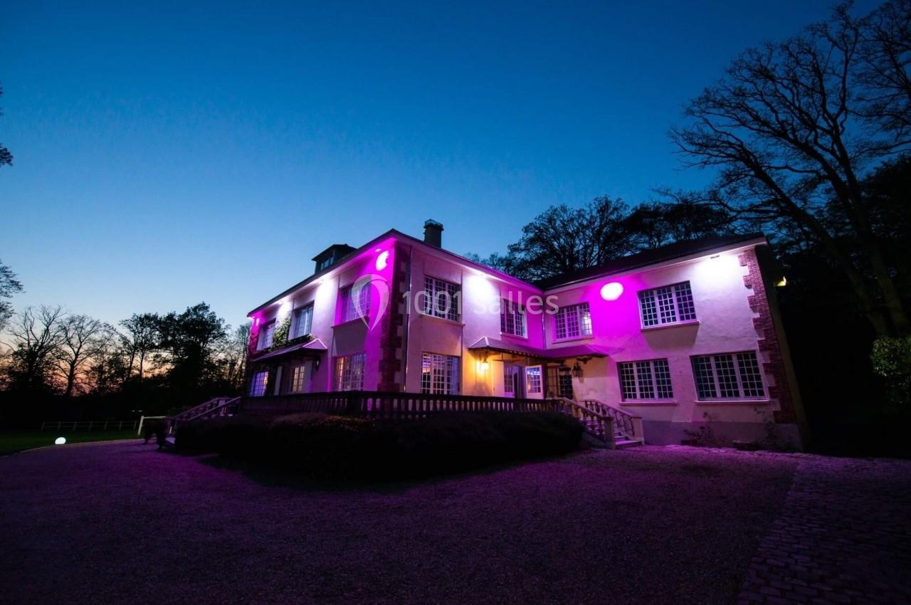 Façade d'une maison éclairée par des lumières violettes au crépuscule, entourée d'arbres et d'un chemin gravillonné.