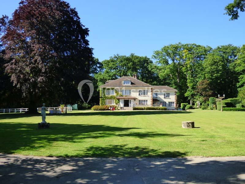 Grande maison entourée d'un vaste jardin arboré sous un ciel bleu clair.