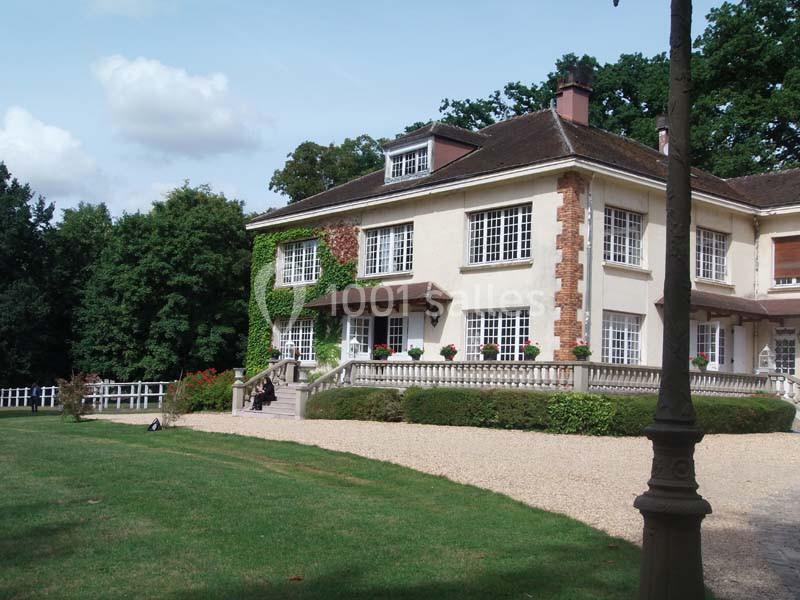 Grande maison avec façade en briques et crépi, entourée de pelouse, arbres et allée gravillonnée.