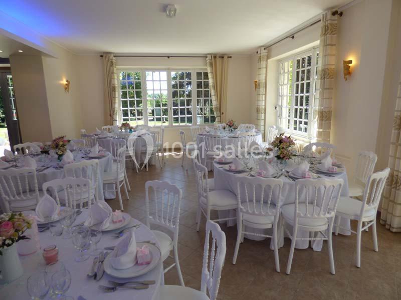 Salle de réception lumineuse avec tables rondes dressées, nappes blanches et décorations florales.