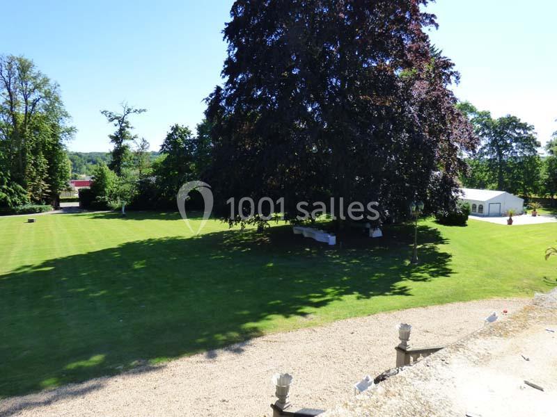 Pelouse verdoyante avec un grand arbre au centre, entourée de végétation et d'un bâtiment blanc en arrière-plan.