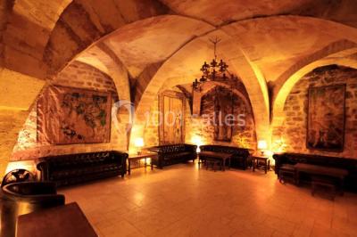 Salle voûtée en pierre éclairée par des lampes et un lustre, avec une table dressée pour un repas.
