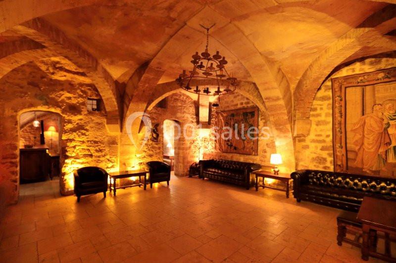 Salle voûtée en pierre avec canapés en cuir, tapisseries murales, éclairage chaleureux et lustre central.