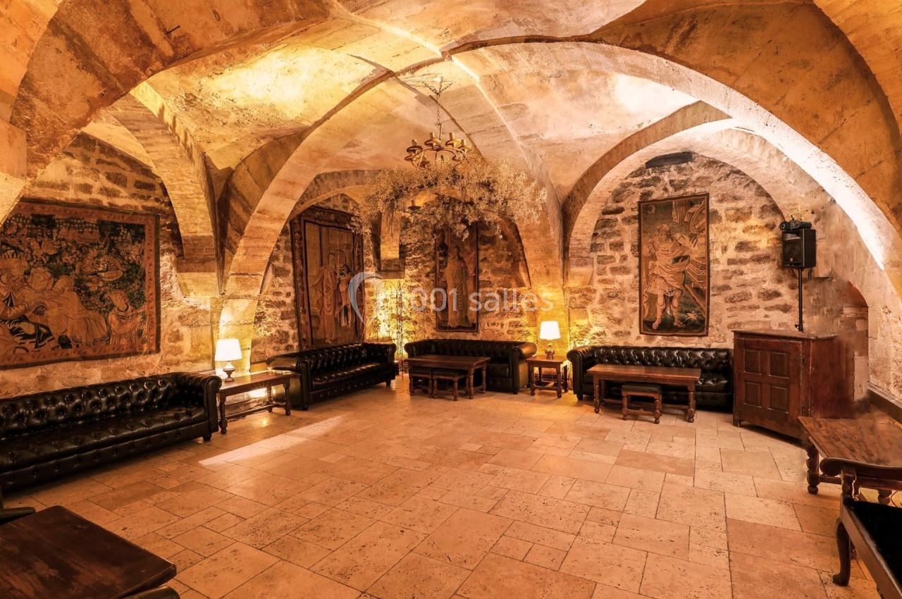 Salle voûtée en pierre avec canapés en cuir, tapisseries murales et éclairage chaleureux.