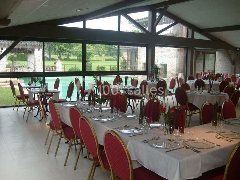 Salle de réception lumineuse avec tables dressées, chaises rouges et vue sur un jardin à travers de grandes baies vitrées.