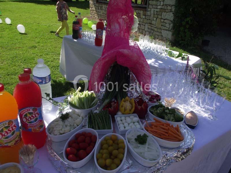 Plateau de légumes frais et condiments disposés sur une table en extérieur, avec boissons et verres en arrière-plan.