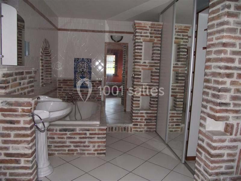 Salle de bain avec murs en briques apparentes, lavabo sur pied, baignoire et carrelage au sol.