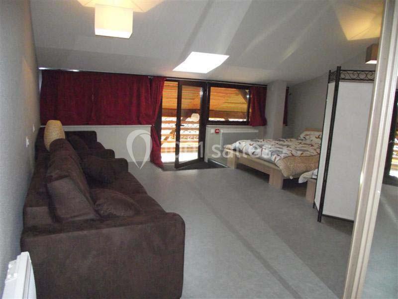 Chambre mansardée avec canapé, lit double, rideaux rouges et accès à un balcon en bois.