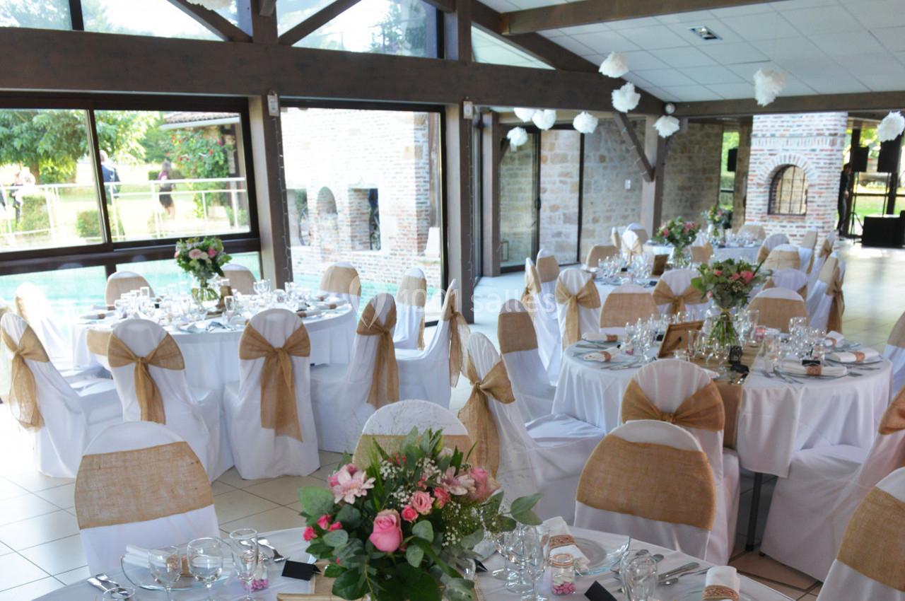 Salle de réception décorée avec des tables rondes, nappes blanches, chaises ornées de nœuds en toile de jute et fleurs.