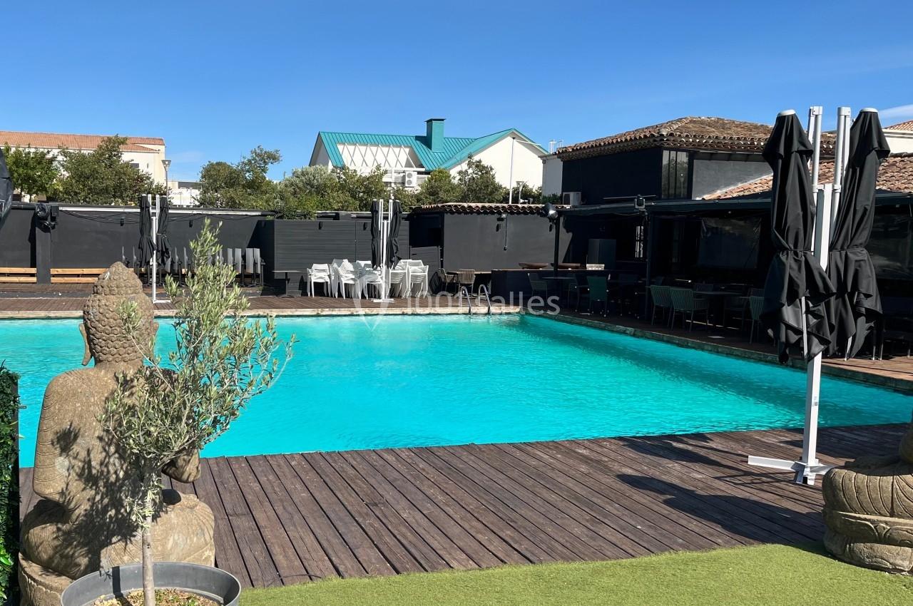 Piscine extérieure entourée de terrasses en bois, statues décoratives et parasols fermés sous un ciel dégagé.