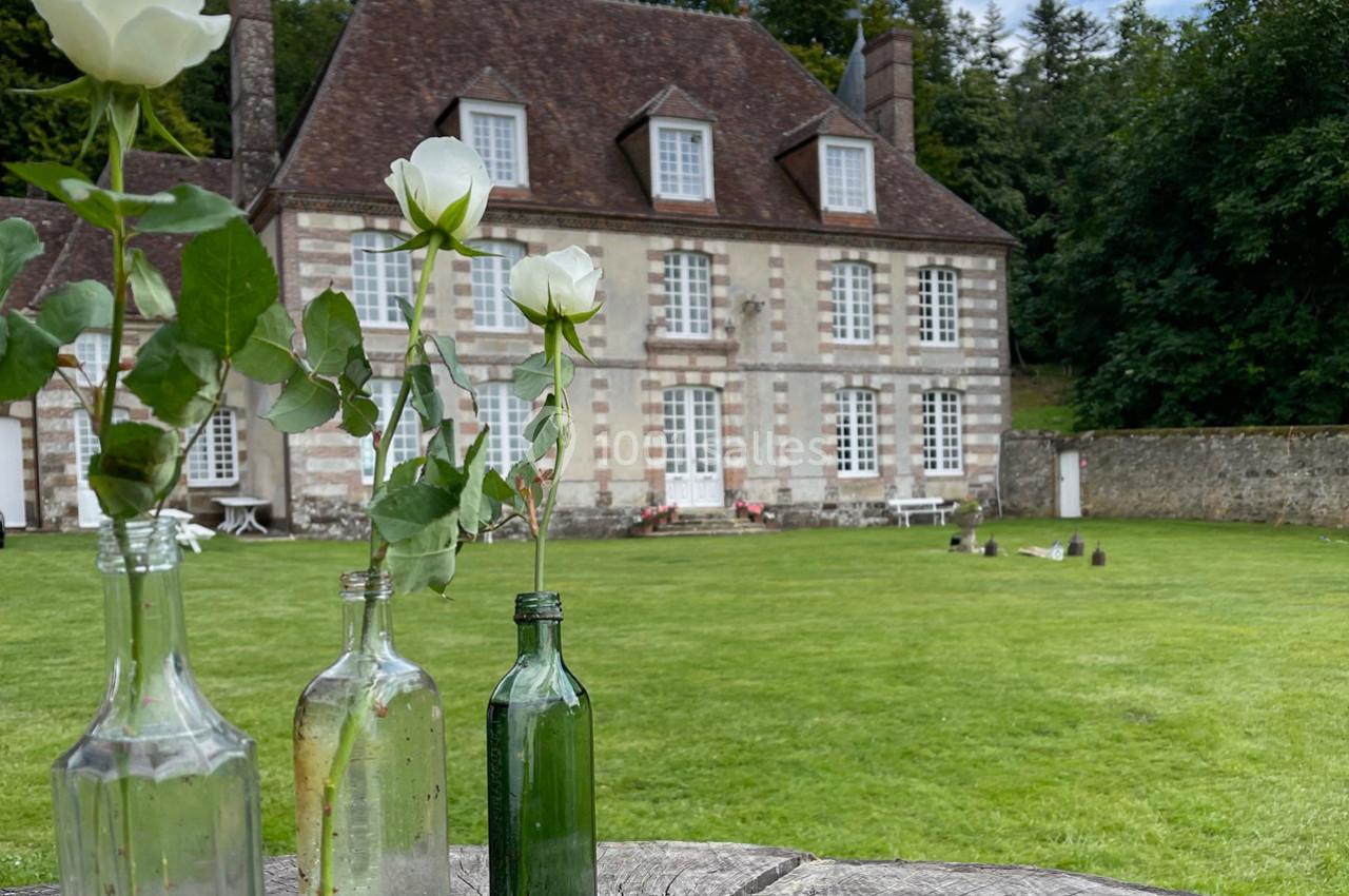 Roses blanches dans des bouteilles en verre devant une maison en pierre avec un grand jardin verdoyant.