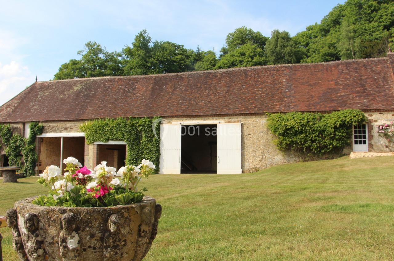 Bâtiment en pierre avec toit en tuiles, portes blanches ouvertes, entouré de pelouse et de végétation.
